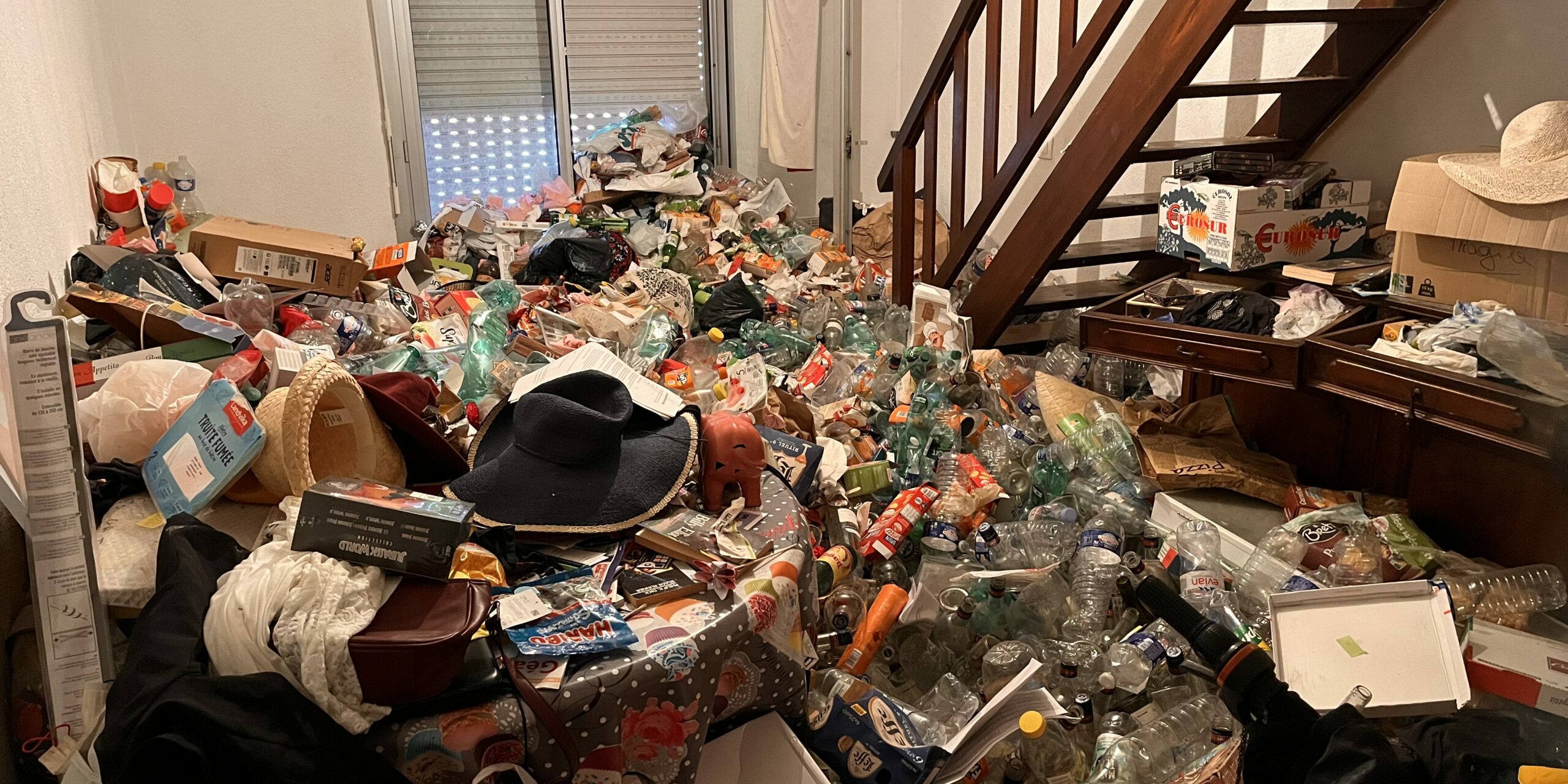 Pièce de vie avec une accumulation d’objets autour du mobilier et d’un escalier en bois.